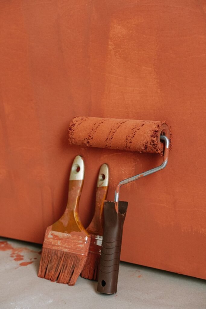 Paintbrushes and a paint roller covered in orange paint leaning on a painted wall